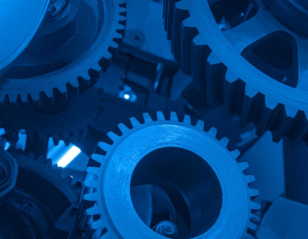 青い光の中で複雑に噛み合う金属製の歯車（ギア）のクローズアップ。AIの仕組みやアルゴリズムが、多くの要素が連携して動くシステムであることを象徴している。