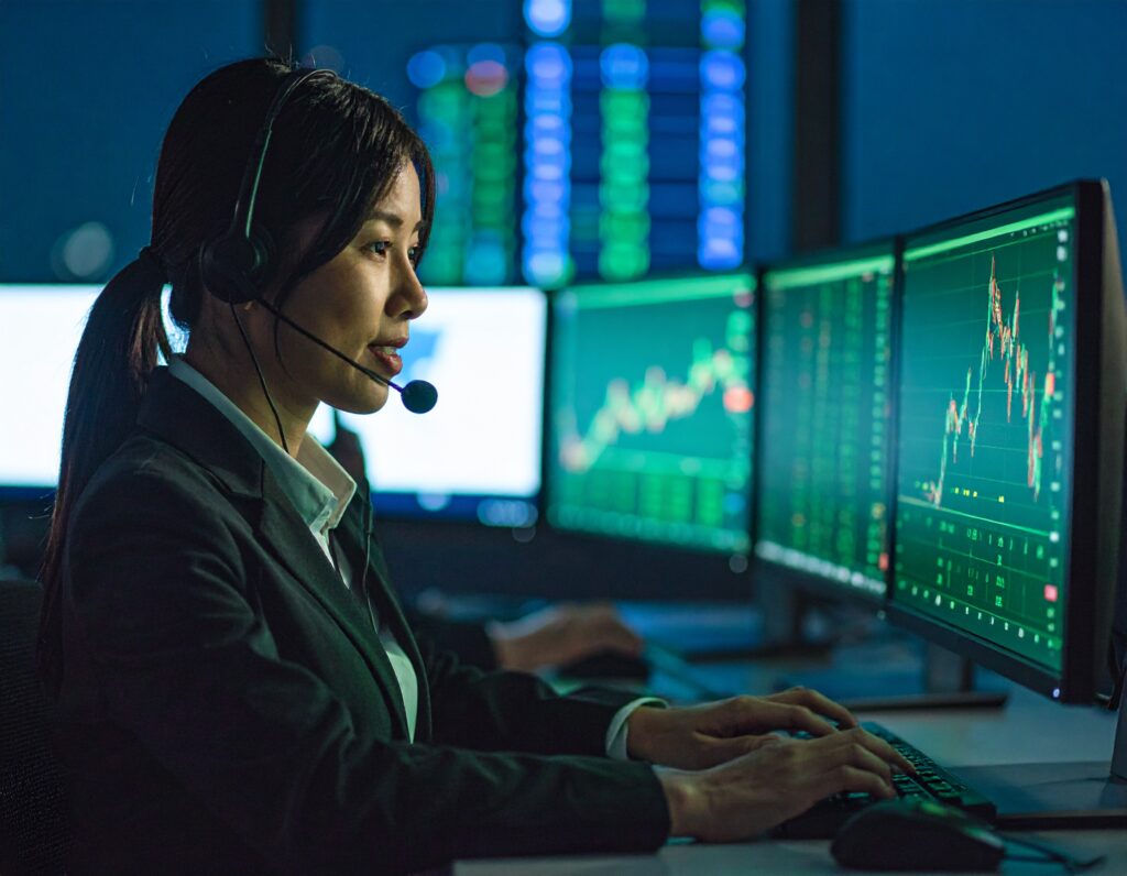 ヘッドセットをつけ、複数のモニターに表示された株価のグラフやデータを見ながら、キーボードを操作する女性オペレーターの側面からの写真。