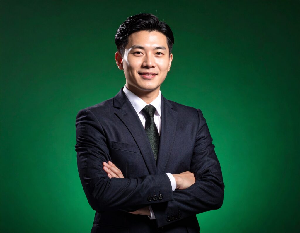 緑色の背景をバックに、スーツ姿で腕を組んで自信のある表情で微笑む、若いアジア系のビジネスマン。