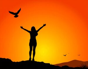 オレンジ色の夕日に向かって、丘の上で両手を大きく広げている人物のシルエット。空には鳥が飛んでいる。